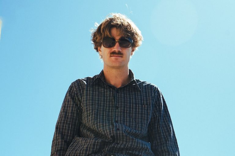 Promotional low-angle portrait of indie artist Coco Elise wearing sunglasses against a clear blue sky.